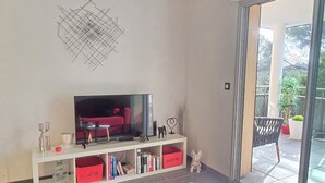 A modern living room with a flat-screen TV, a white entertainment unit with books, and a glass door leading to a balcony with potted plants. - Saint-Raphaël: Apartment Near Beach & Pool (Saint-Raphaël)