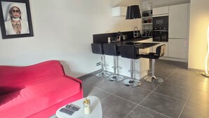 A modern living room with a red sofa, a small coffee table, and a kitchen area with a bar stool seating. - Saint-Raphaël: Apartment Near Beach & Pool (Saint-Raphaël)