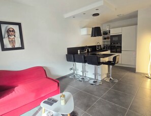 A modern living room with a red sofa, a small coffee table, and a kitchen area with a bar stool seating. - Saint-Raphaël: Apartment Near Beach & Pool (Saint-Raphaël)