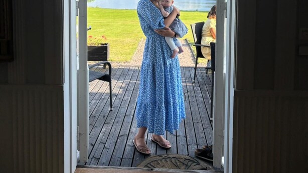 Una mujer con vestido azul sostiene a un bebé en una cubierta de madera con vista al lago.