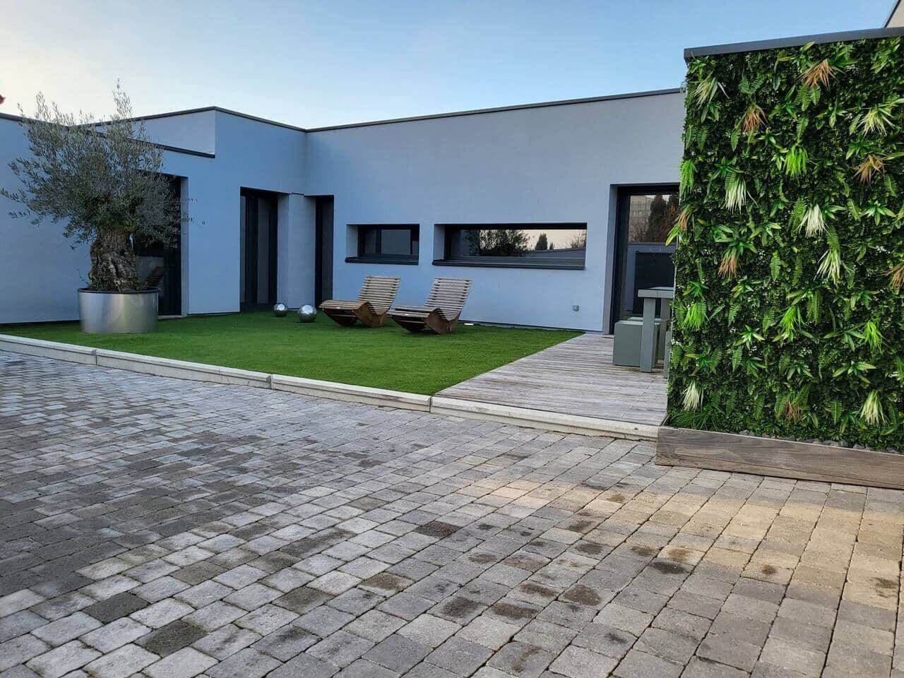Un bâtiment moderne doté d’un toit plat, d’une allée pavée et d’une pelouse bien entretenue, agrémentée de deux chaises en bois.