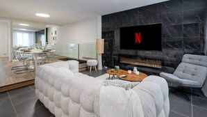 A modern living room with a fireplace, a flat-screen TV mounted on the wall, and a dining area visible through a glass partition. - To complete (Reims)