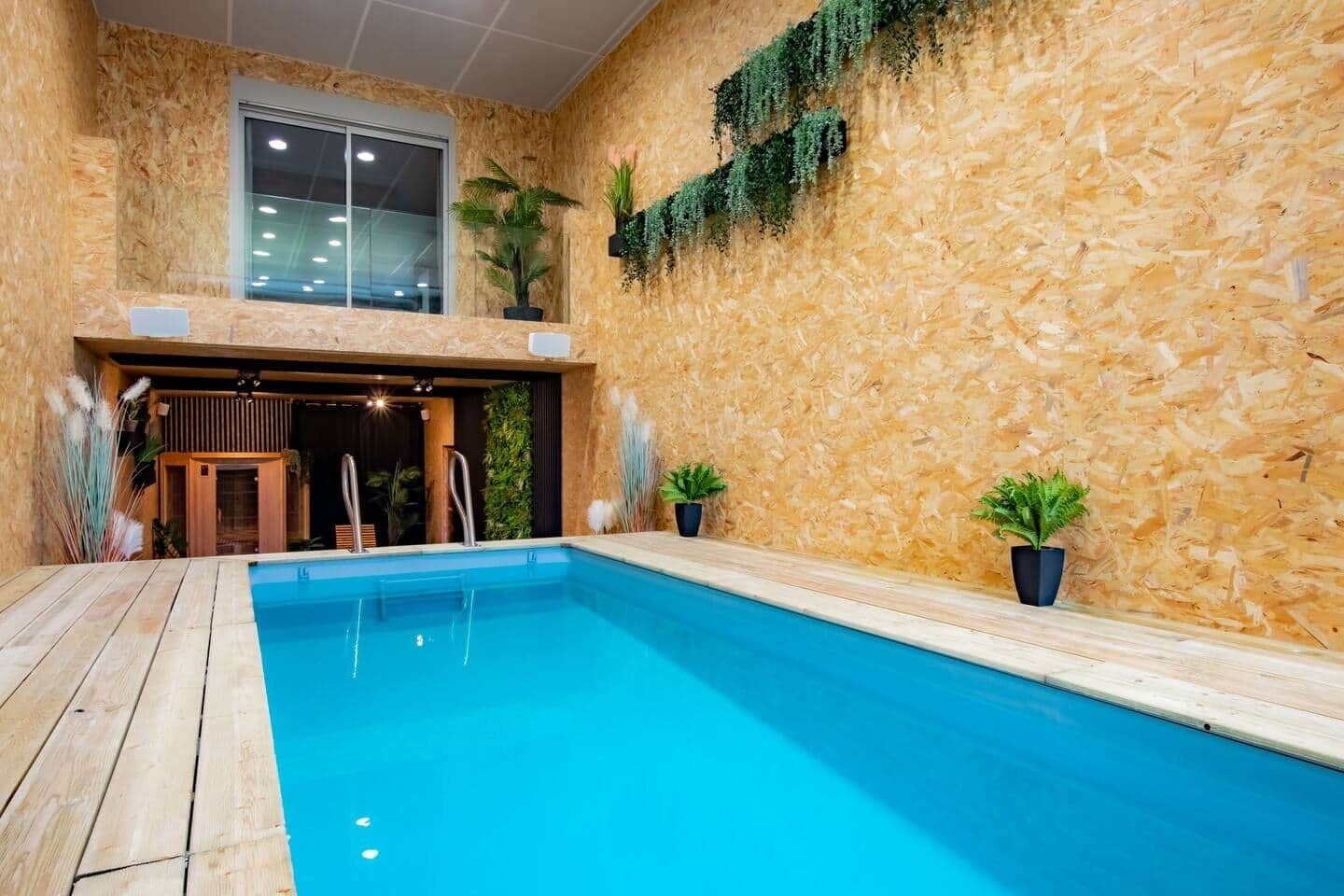 Une piscine intérieure dotée d’une terrasse en bois, d’une entrée avec porte vitrée et de plantes en pot.