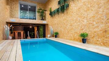 Une piscine intérieure dotée d’une terrasse en bois, d’une entrée avec porte vitrée et de plantes en pot.