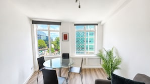 Una habitación moderna con una mesa de vidrio, sillas negras, un sofá gris y un ventanal con vistas a edificios y árboles.