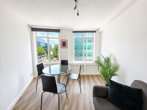Ein modernes Zimmer mit Glastisch, schwarzen Stühlen, einem grauen Sofa und einem großen Fenster mit Blick auf Gebäude und Bäume.