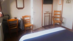 Ein Schlafzimmer mit einem Bett, zwei Holzstühlen, einem Schreibtisch mit Stuhl, einem Kleiderständer, einem Fernseher und einer Tür.