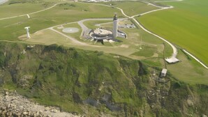 Vista aérea de una zona costera con un faro destacado, campos verdes y una playa.