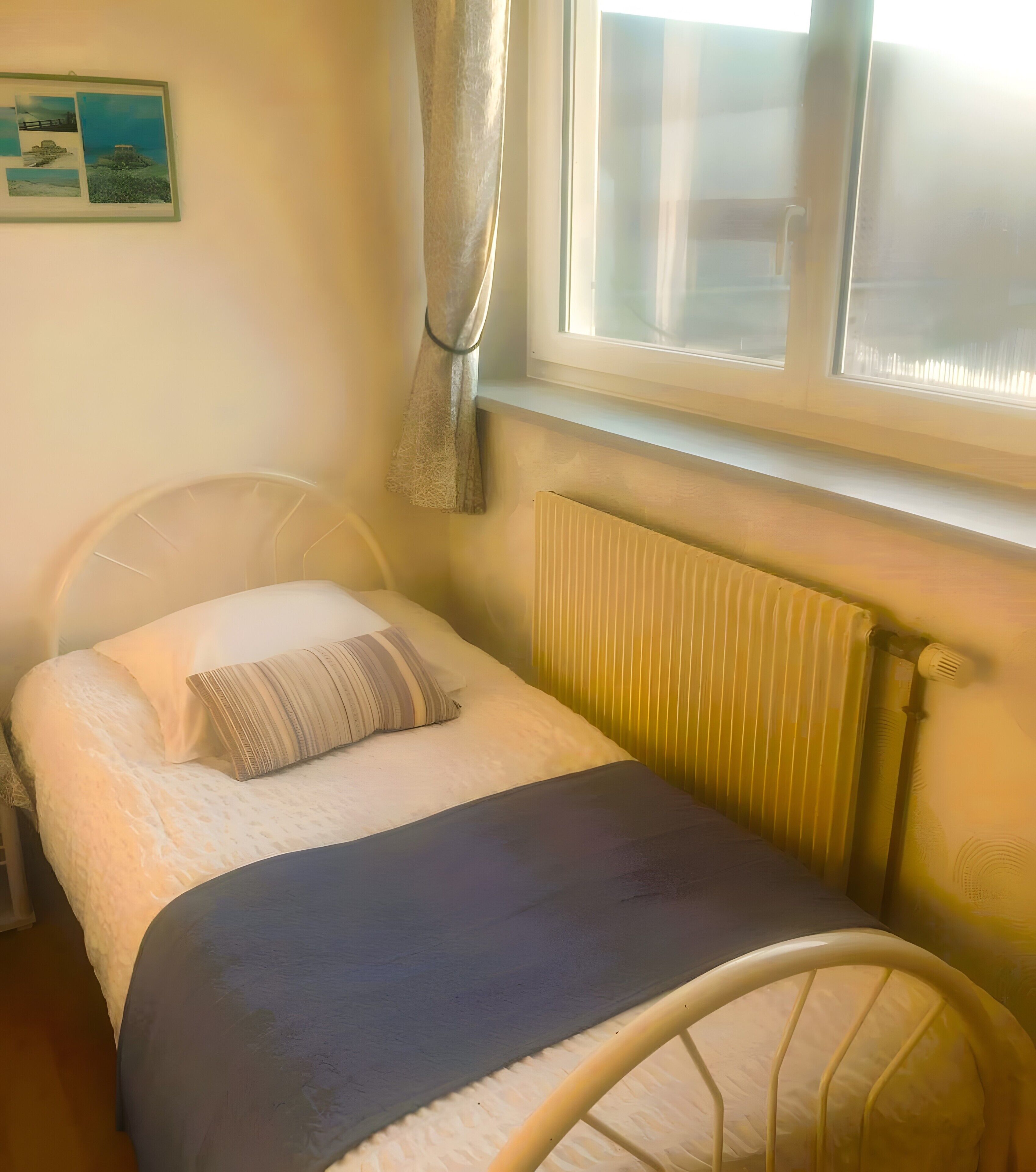 A single bed with a headboard, a window with curtains, a radiator, and a wall-mounted picture.