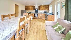 A compact kitchen with wooden cabinets, a dining area with a table and chairs, and a sofa with a green pillow. - Host & Stay | Morley Cottage (Devon)
