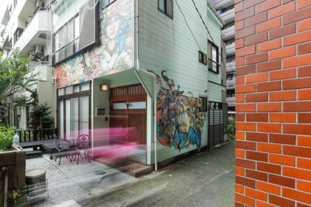 Un edificio con un mural colorido en la pared exterior, un pequeño espacio al aire libre con mesas y sillas, y una pared de ladrillo a la derecha.