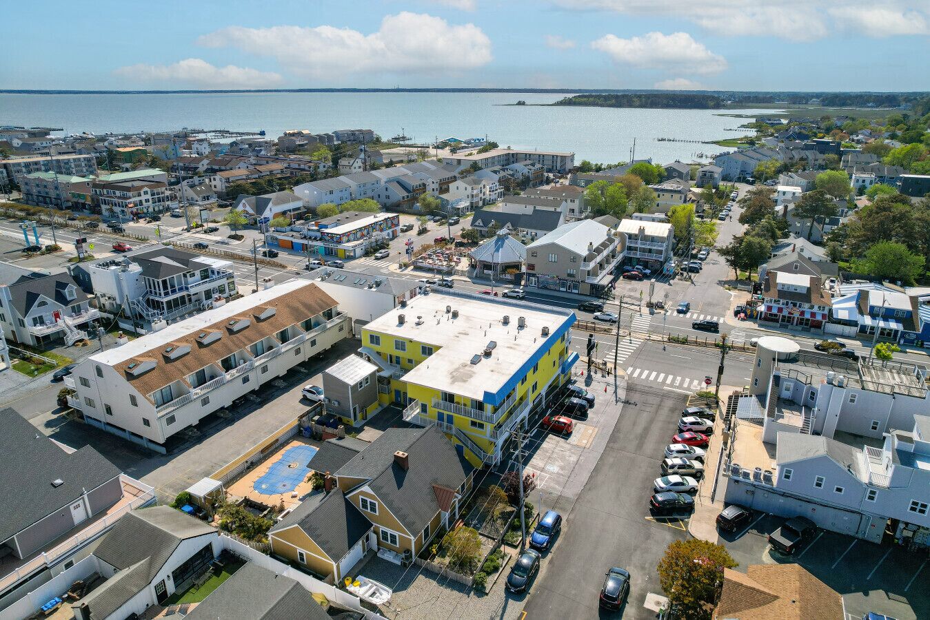 An aerial view of a coastal town with a mix of residential and commercial buildings, a marina, and a clear view of the sea.