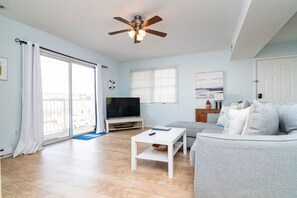 Una sala de estar con sofá, televisor, ventilador de techo y balcón con cortinas.
