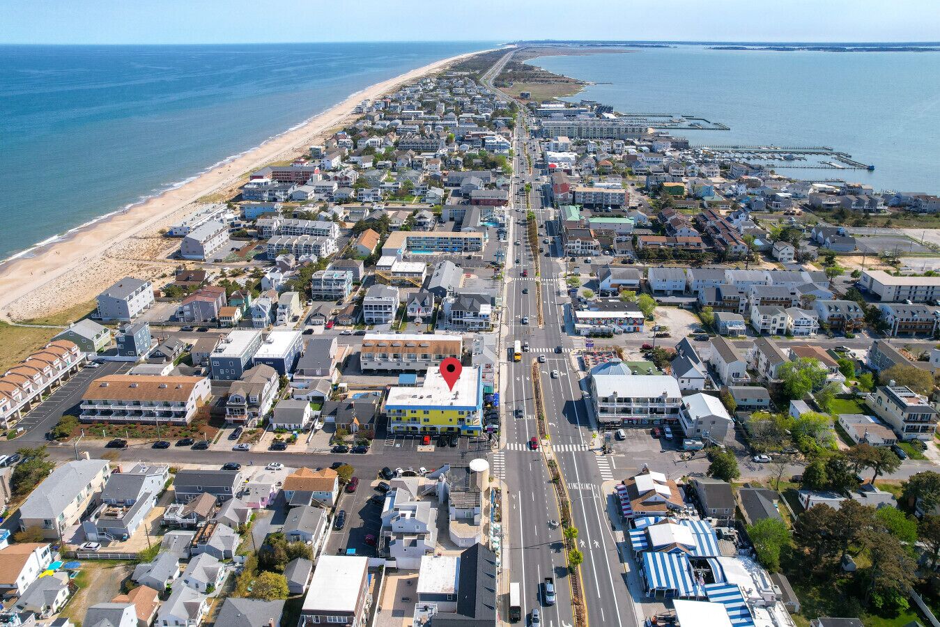 An aerial view of a coastal town with a long beachfront, a central road, and a prominent building with a red roof.
