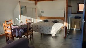Een slaapkamer met een bed, een bureau met stoel, een klein tafeltje met een plant en uitzicht op een keuken.