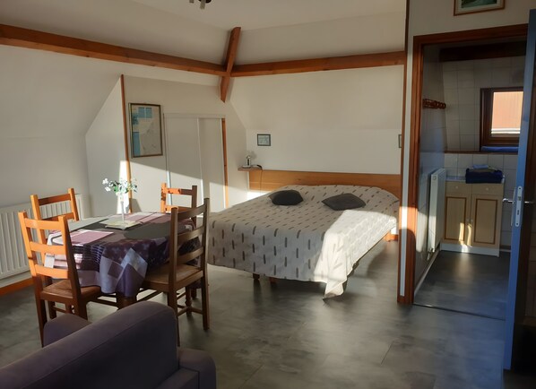 Een slaapkamer met een bed, een bureau met stoel, een klein tafeltje met een plant en uitzicht op een keuken.