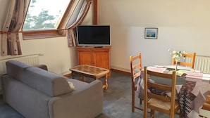 Een woonkamer met een bank, een tv op een houten kast, een eettafel met stoelen, en een raam met gordijnen.