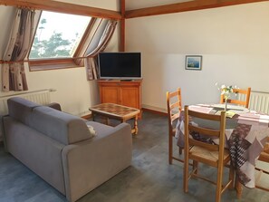 Ein Wohnzimmer mit Sofa, Fernseher auf einer Holzkommode, Esstisch mit Stühlen und einem Fenster mit Vorhängen.
