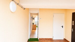 Um corredor com uma porta branca, uma placa de número e uma área de recepção visível através de uma porta aberta.