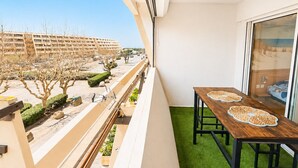 Uma varanda com mesa e cadeiras de madeira, vista para um estacionamento e edifícios, além de uma área com grama sintética verde.