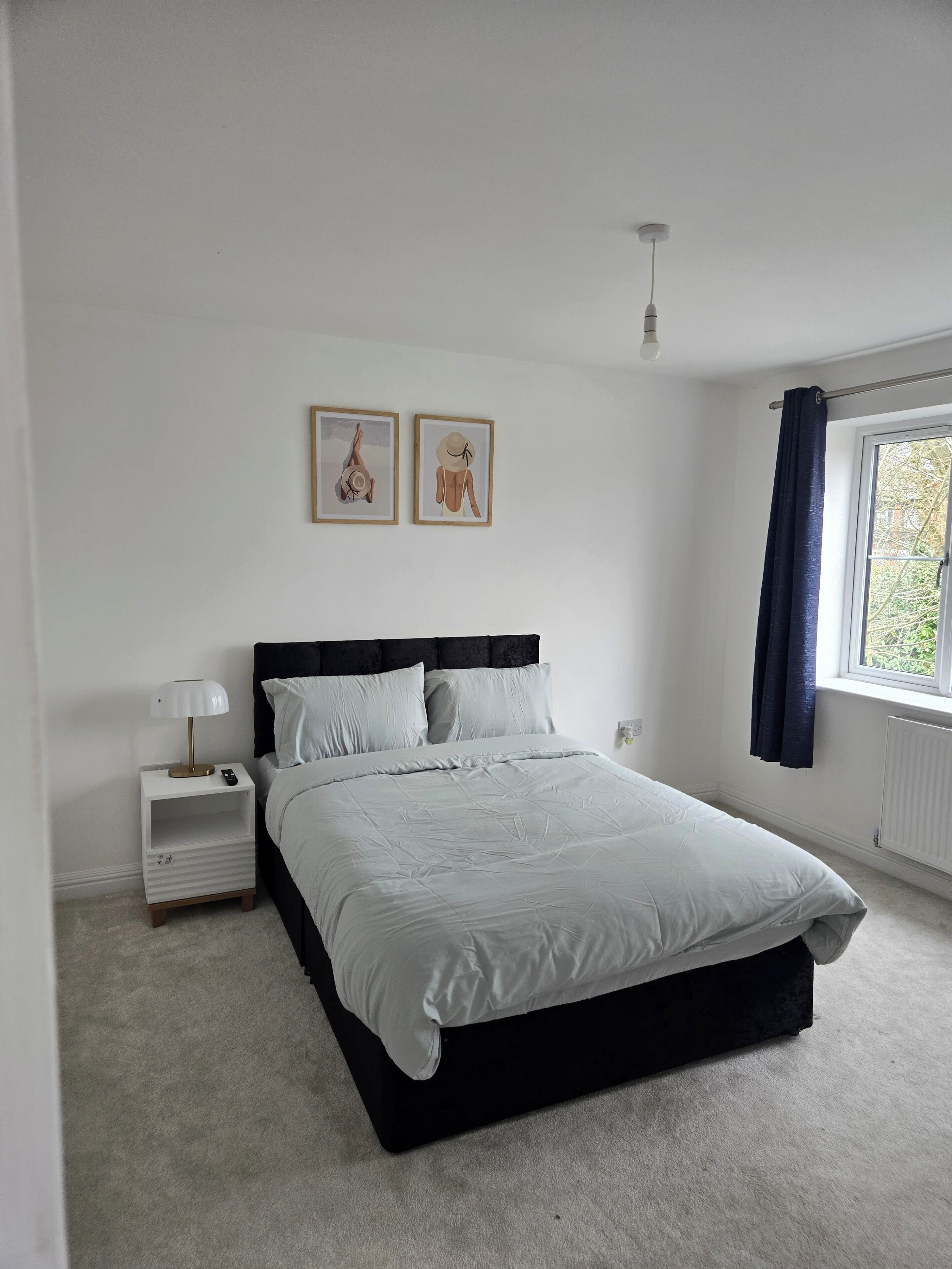 A bedroom with a bed, a nightstand, a lamp, two framed pictures on the wall, and a window with curtains.