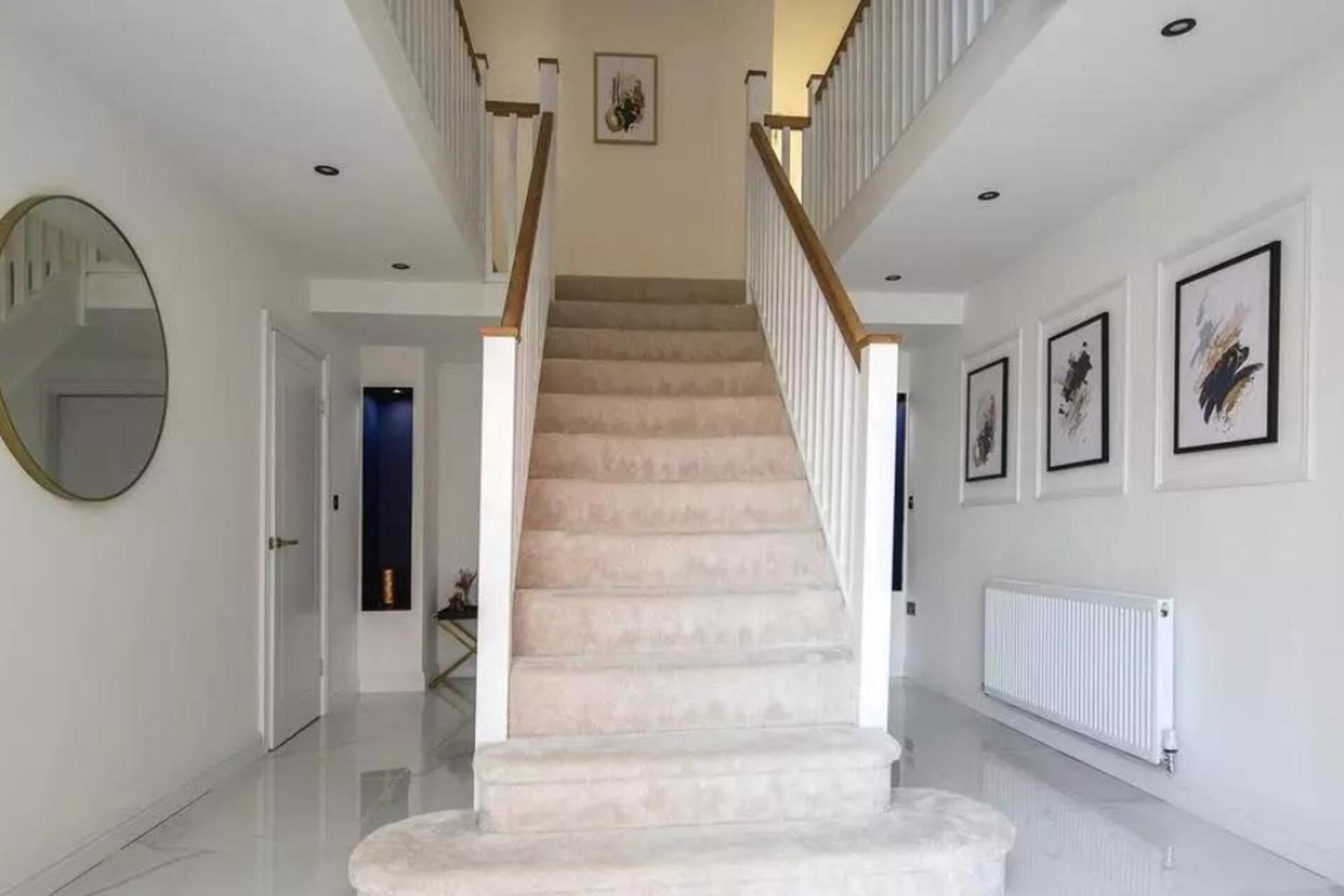 A staircase with a wooden handrail and beige carpet, leading to a landing with a mirror and framed artwork on the walls.
