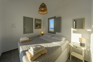 A bedroom with a bed, bedside tables, a lamp, a hanging light, a painting, and a view of the sea. - Villa Meloria, Divine Sea Views, Private Pool, Furnished Terrace, Finikia (Santorini)