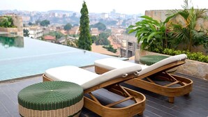 A rooftop lounge with two wooden lounge chairs and a woven ottoman, overlooking a cityscape and a swimming pool.