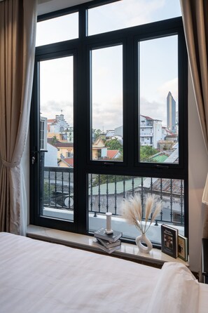 A bedroom with a large window, a bed, a bedside table with books and a vase, and a view of buildings outside. - An Dinh La Maison by Teky House (Thuan Hoa)