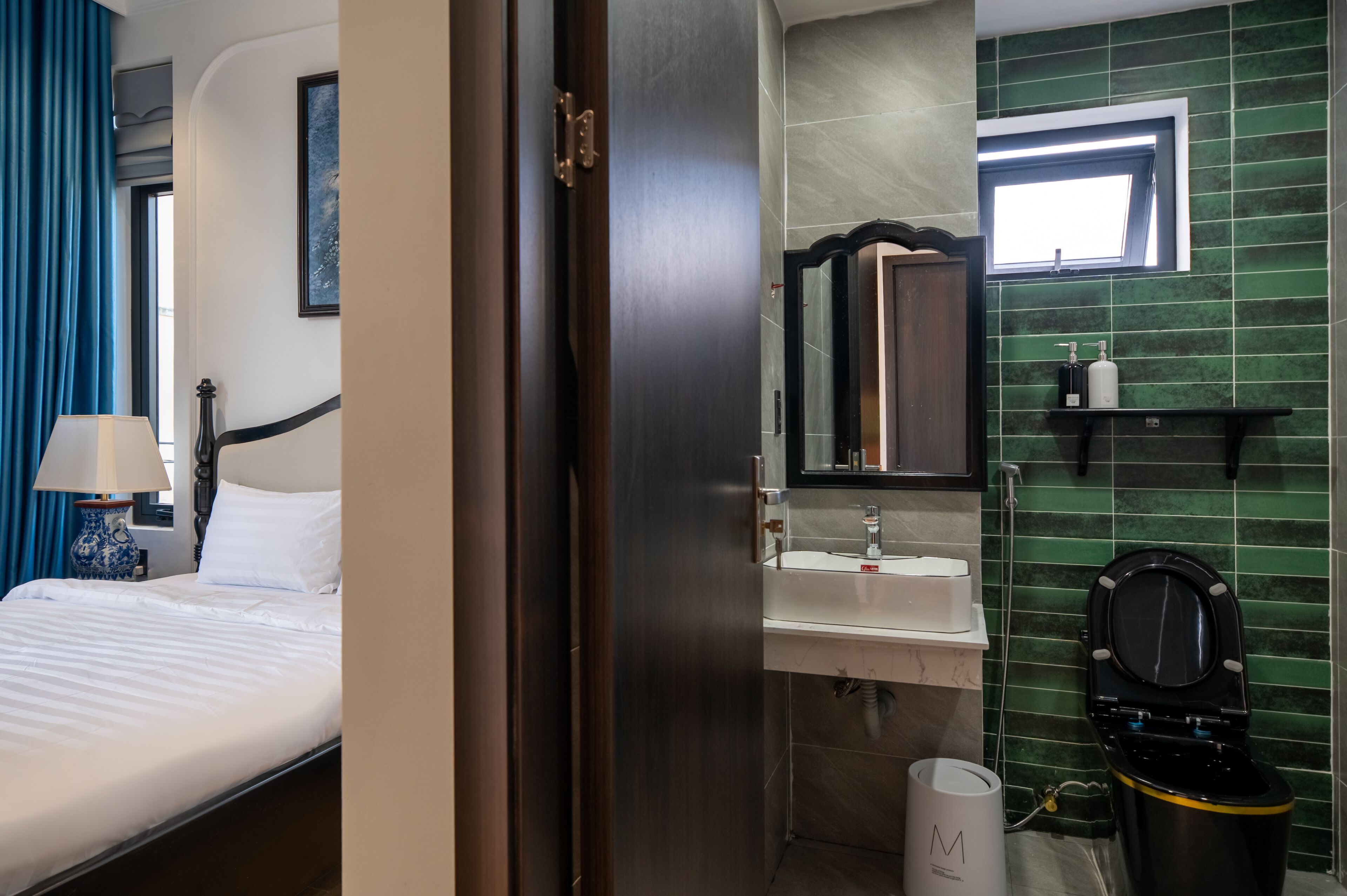 A hotel room with a bed, a nightstand, a lamp, a bathroom with a sink, mirror, toilet, and green tiled walls.