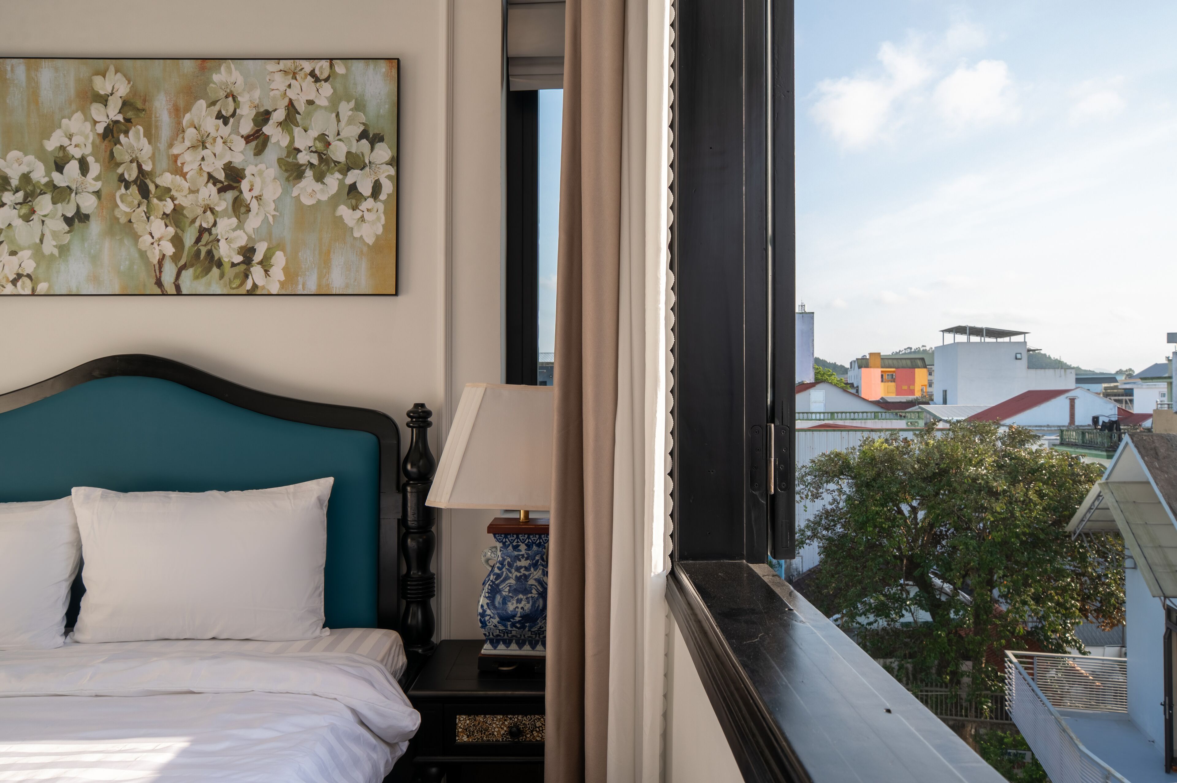 Quarto de hotel com cama, quadro, mesa de cabeceira, luminária e vista para edifícios e árvores.