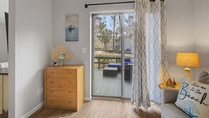 Una stanza con una porta scorrevole in vetro che dà su una terrazza esterna, un settimino in legno, un divano con un cuscino e una lampada.