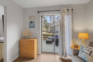 Een kamer met een schuifglasdeur die uitkomt op een terras, een houten dressoir, een bank met een kussen en een lamp.