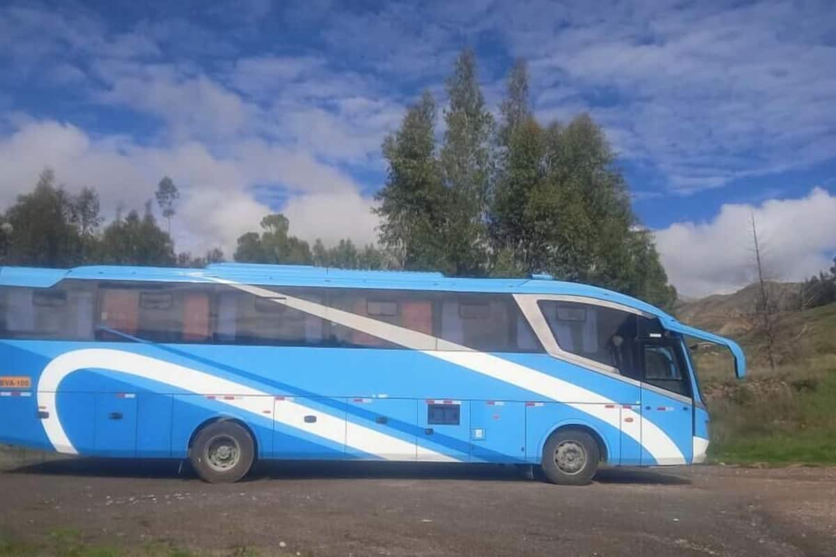 A large blue and white bus parked on a dirt road with trees and hills in the background.