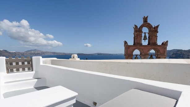 Un balcón blanco con escalones, una torre campanario y vista al mar y montañas distantes.