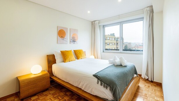 A modern hotel room with a large bed, a bedside table, a nightstand, a window with a view of buildings, and two wall art pieces. - GuestReady - Charming Braga retreat (Braga)