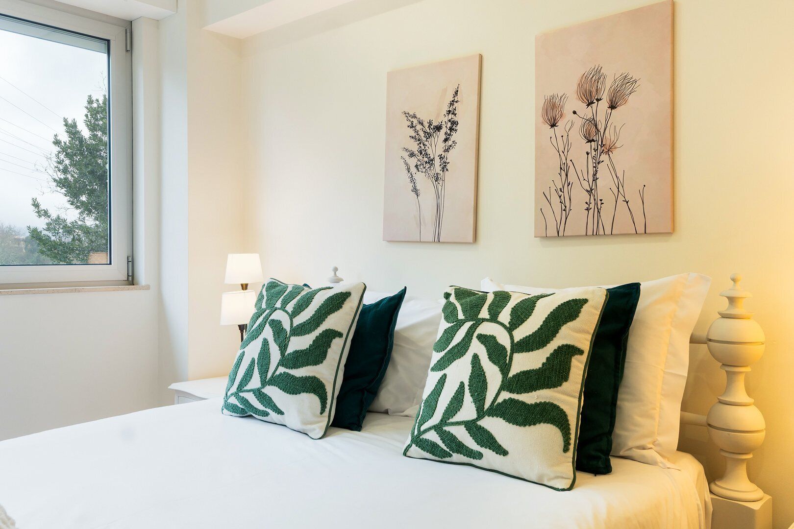 A bedroom with a bed, two green leaf-patterned pillows, a nightstand, a lamp, and two wall-mounted floral artwork pieces.