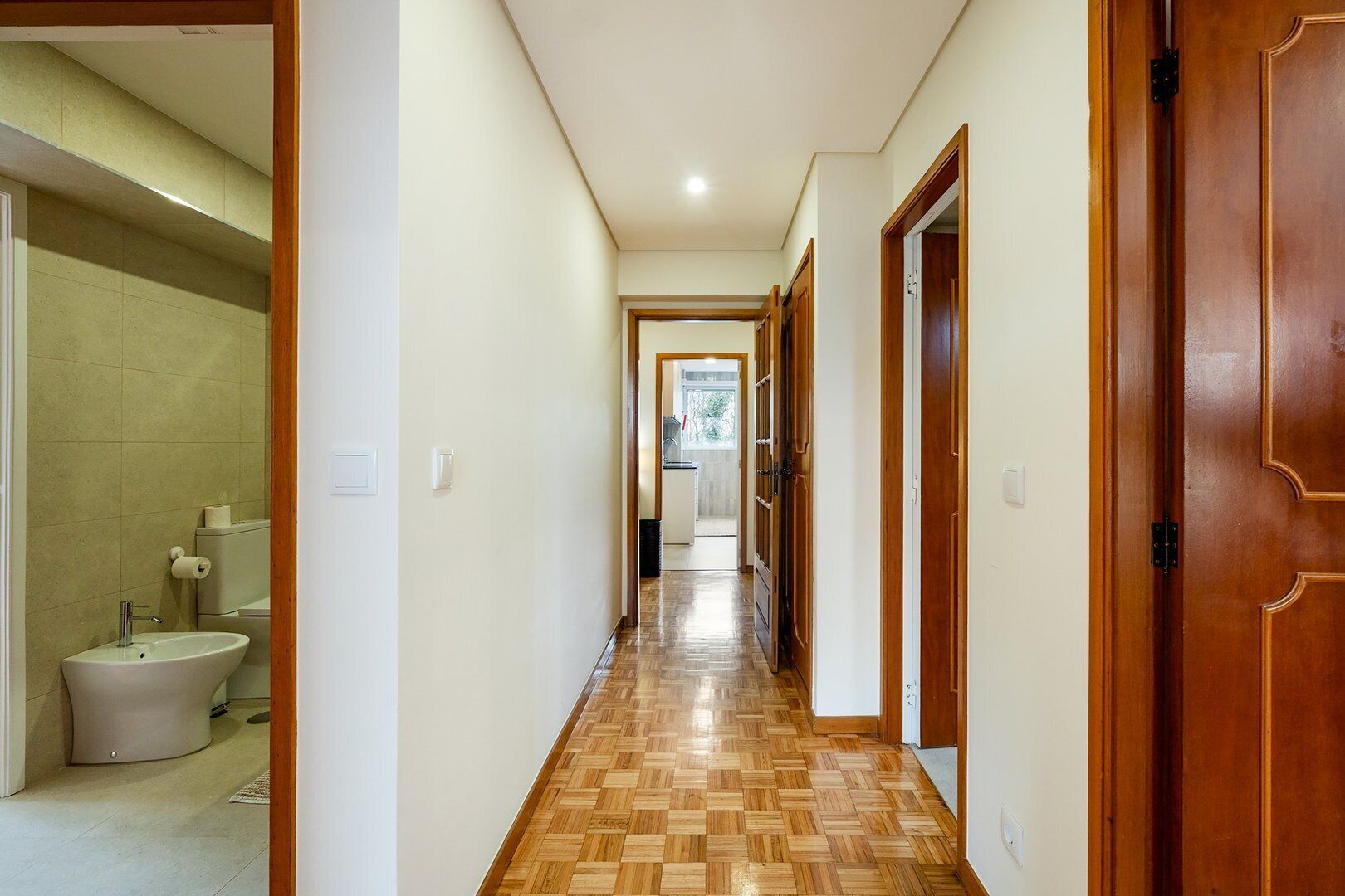 Un pasillo con puertas de madera, un baño con baldosas y un suelo con estampado tipo espiga.