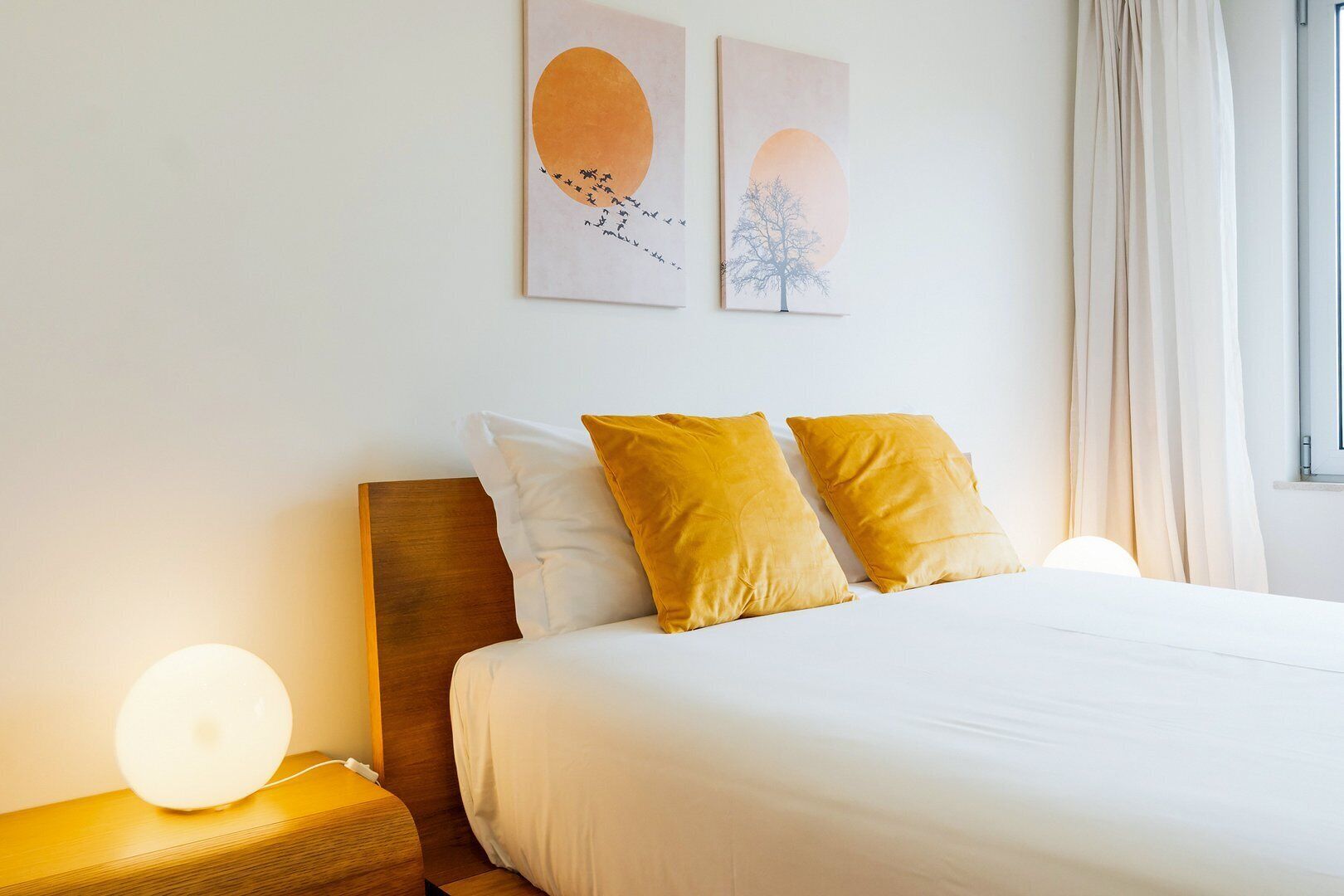 A neatly arranged hotel room with a large bed, two pillows, a bedside table with a lamp, and two wall art pieces depicting birds and a tree.