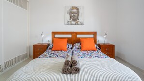 Une chambre à coucher avec un lit, deux tables de chevet, une tête de lit, une œuvre d’art accrochée au mur et une serviette roulée posée sur le lit.