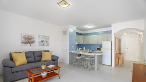 Un salon moderne avec un canapé gris, une table basse en bois et un coin cuisine doté d’un réfrigérateur blanc et de carreaux bleus.