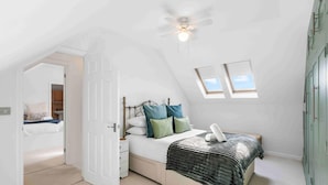 A bedroom with a sloped ceiling, a bed with a white headboard, two bedside tables, a nightstand, a ceiling fan, and two windows. - Sea Palm By My Getaways (Lancing)