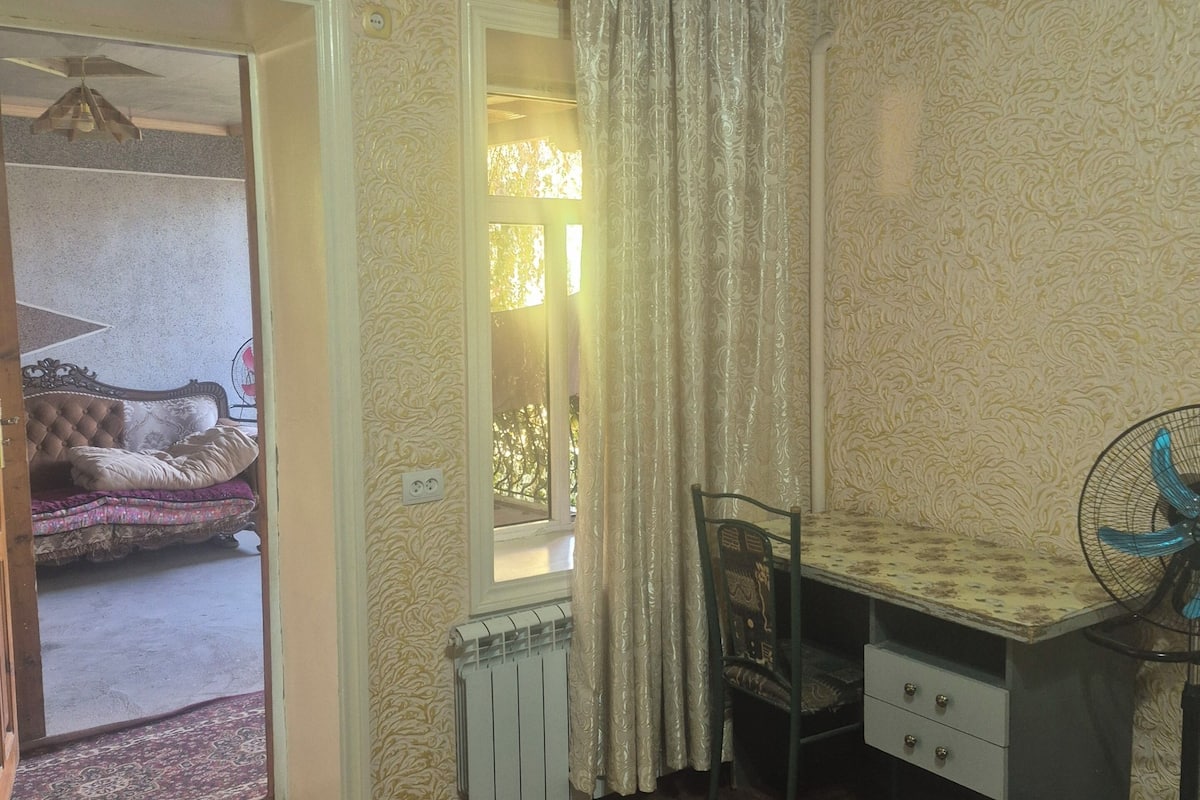 A room with a wooden ceiling, a radiator, a window with curtains, a bed with a patterned bedspread, and a desk with a chair.