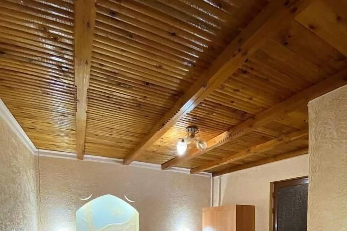 A bedroom with a wooden ceiling, a bed with floral bedding, a wooden bench, and a window with a view.