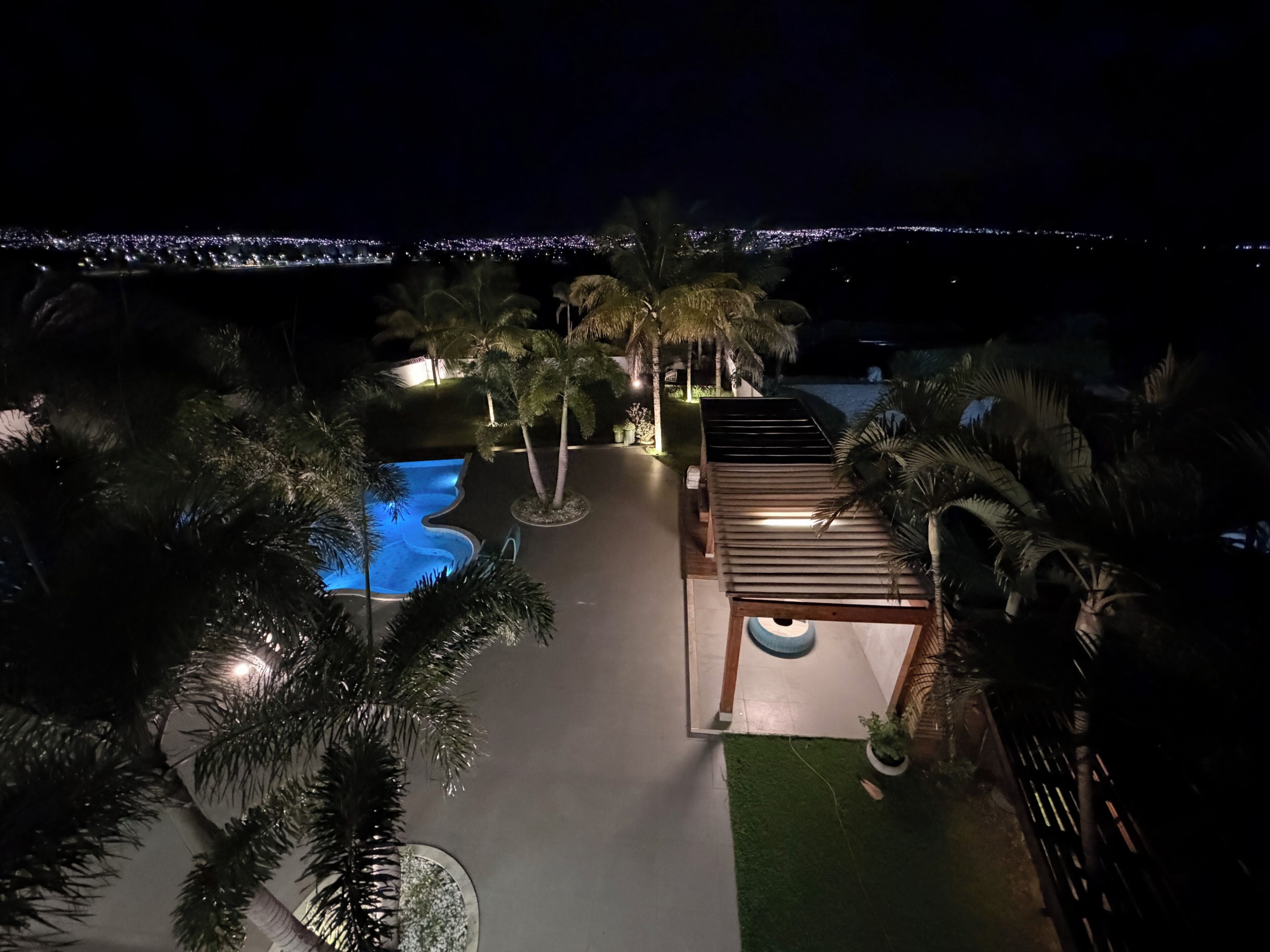 Vista noturna de um resort com piscina, palmeiras e área de estar coberta.