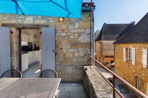 A stone building with a kitchen visible inside, a balcony with a blue awning, and a car parked outside.