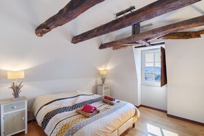 A bedroom with a bed, wooden beams, a window, and a nightstand with a lamp.