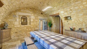 Une pièce aux murs de pierre, avec une longue table, des bancs et une porte donnant sur une autre pièce.