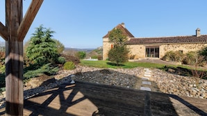 Un bâtiment en pierre avec un toit en tuiles, entouré d’un jardin agrémenté d’une terrasse en bois.