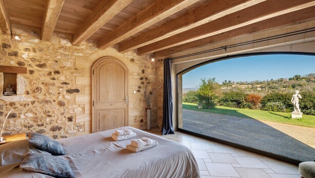 Une chambre avec un mur en pierre, des poutres en bois, un lit avec des serviettes et une vue sur une route et la verdure.